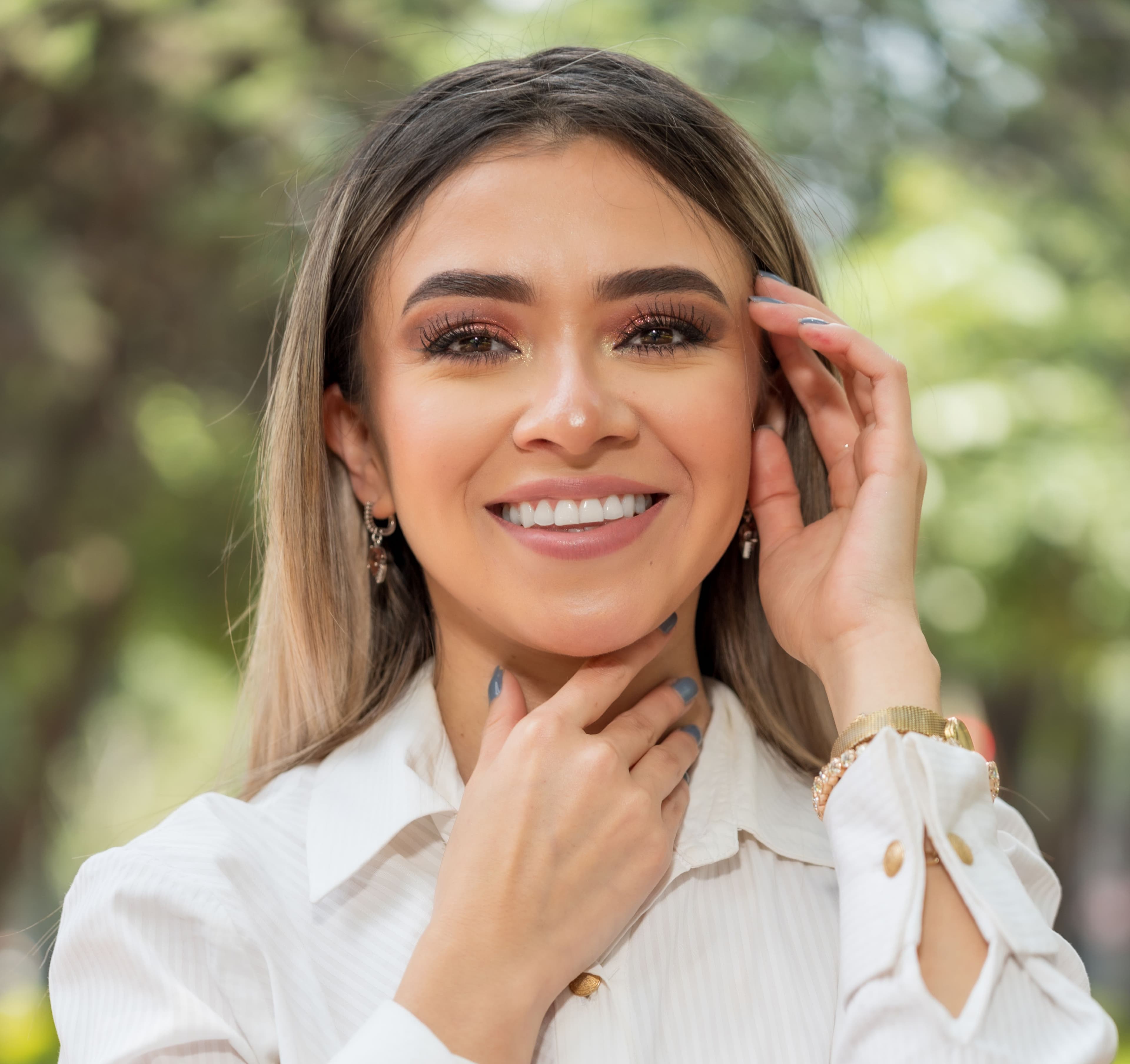 Smiling woman with blonde-highlighted hair and shimmery makeup posing in a white shirt outdoors.
