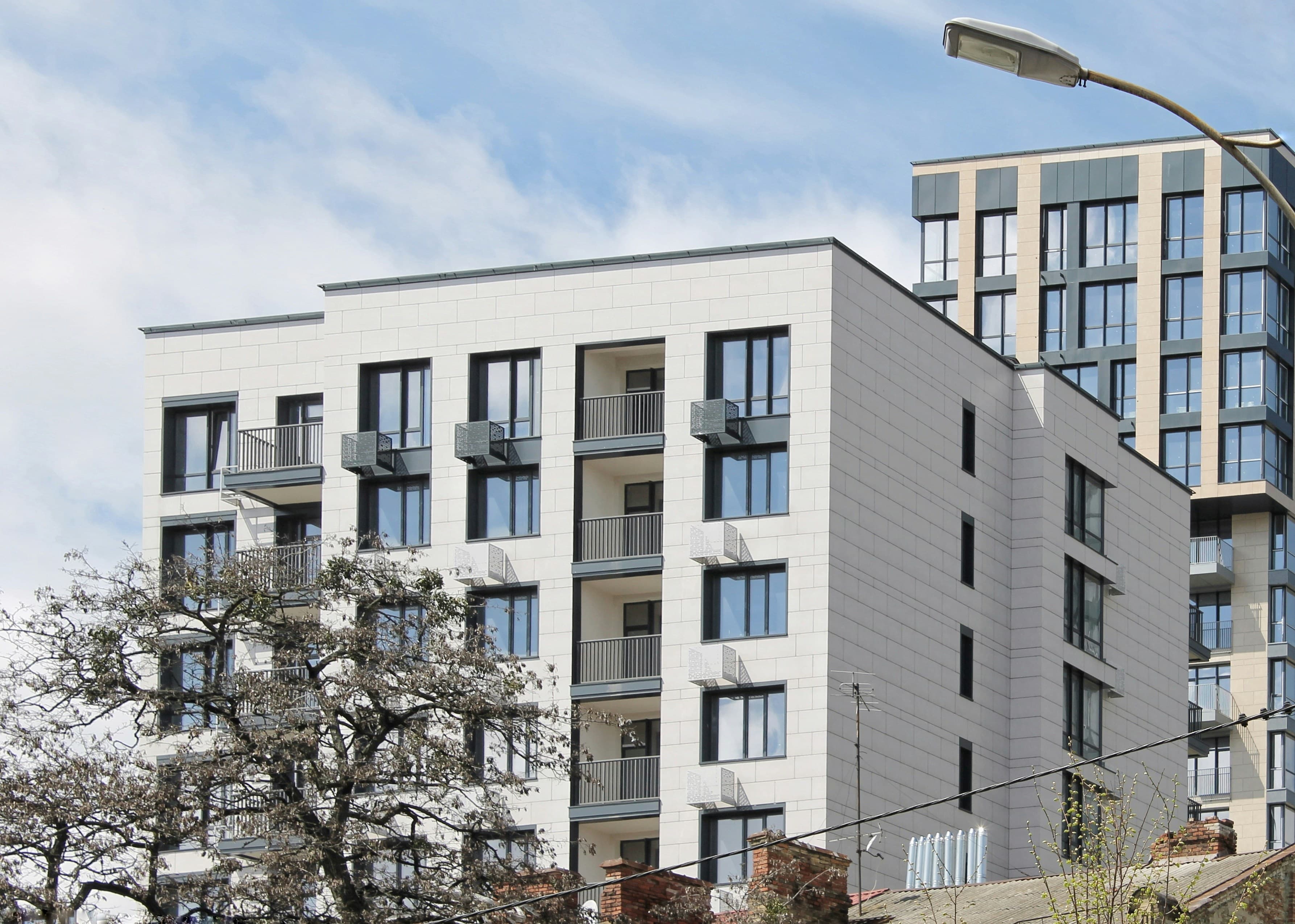 Modern white apartment buildings with balconies and windows under a blue sky and bare tree.