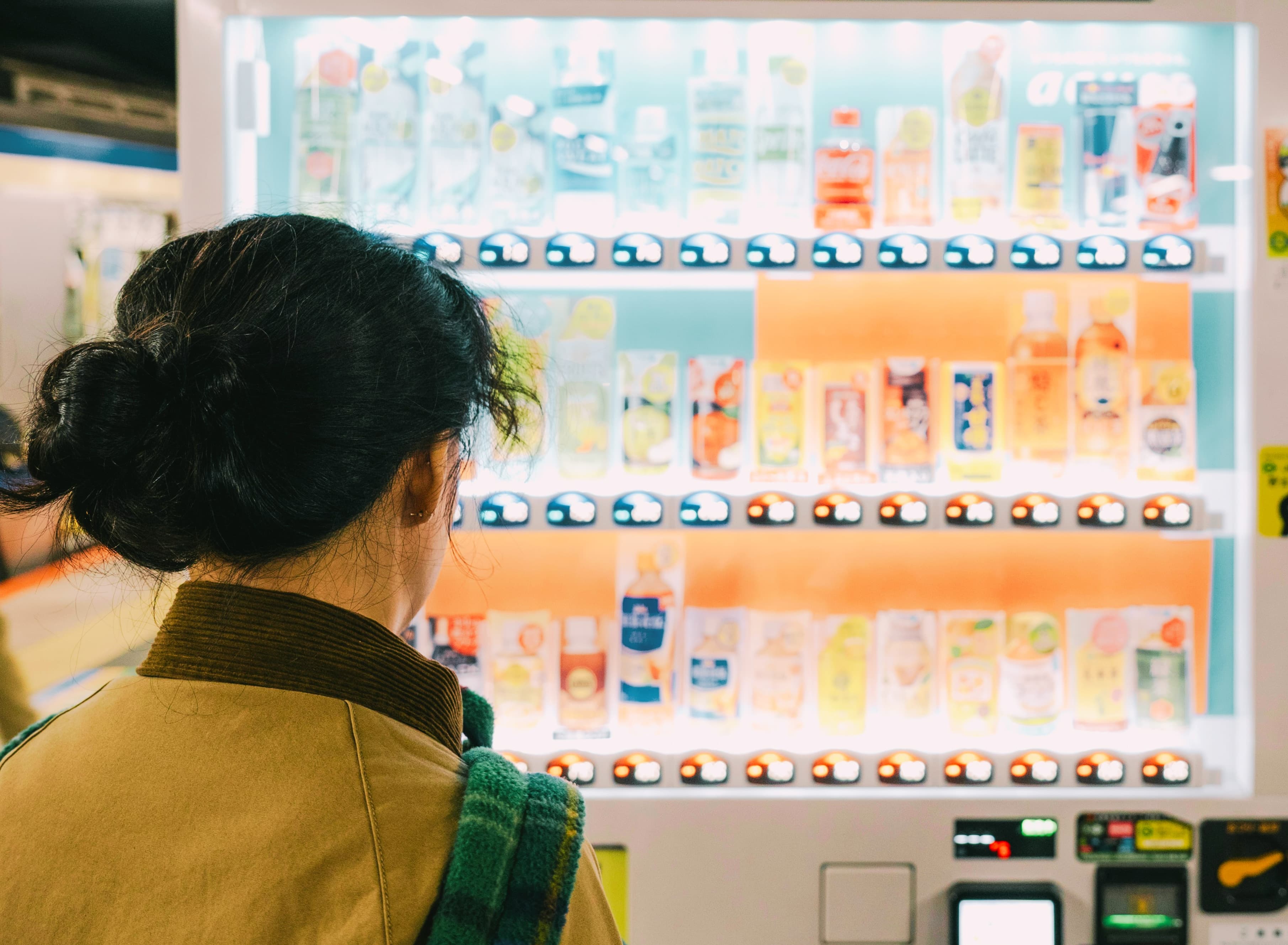 Woman with hair in a bun looking at a brightly lit vending machine with drinks.
