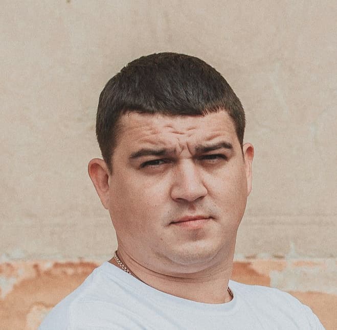A man with short dark hair and a serious expression wearing a white t-shirt.