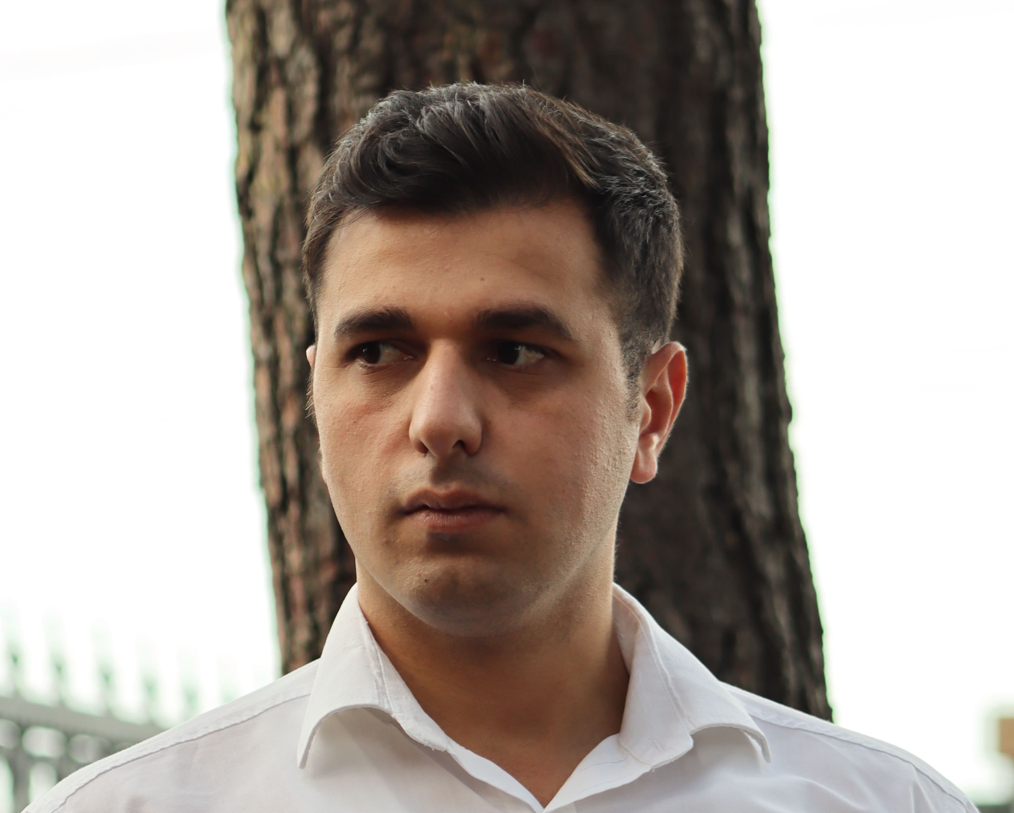 A serious man in a white shirt looking away, standing before a tree trunk.